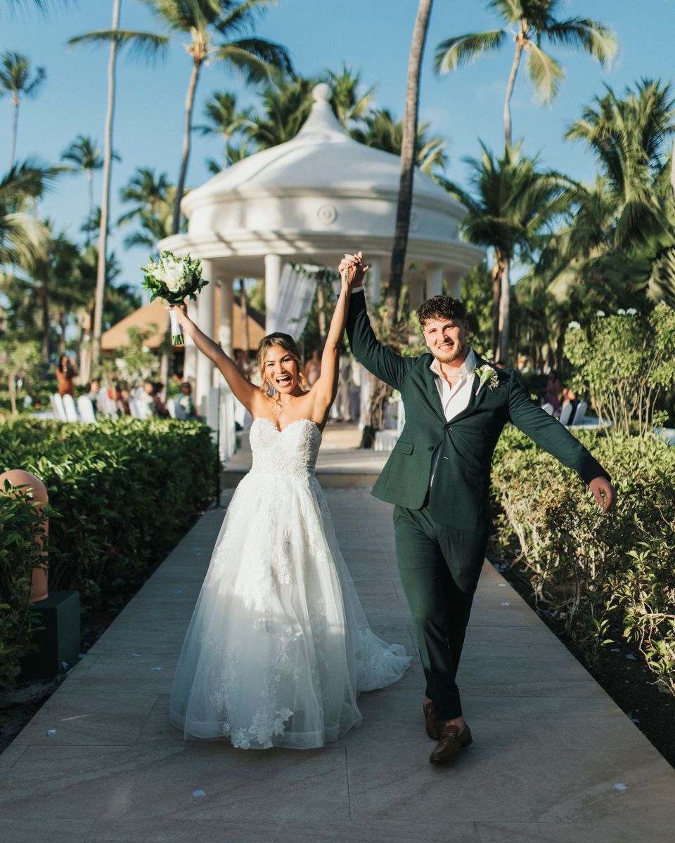 Newlywed couple walking down a garden path after an outdoor destination wedding ceremony with palm trees and a white gazebo in the background.