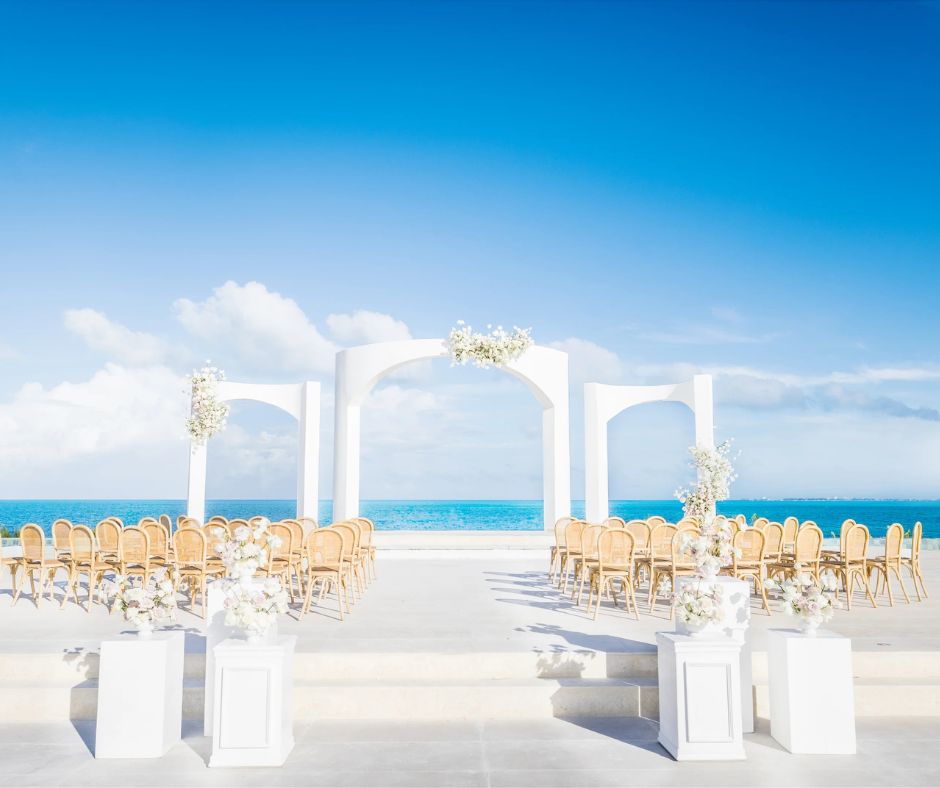 Beachfront destination wedding ceremony setup with white arches, floral arrangements, and rows of wooden chairs overlooking turquoise ocean waters under a clear blue sky.