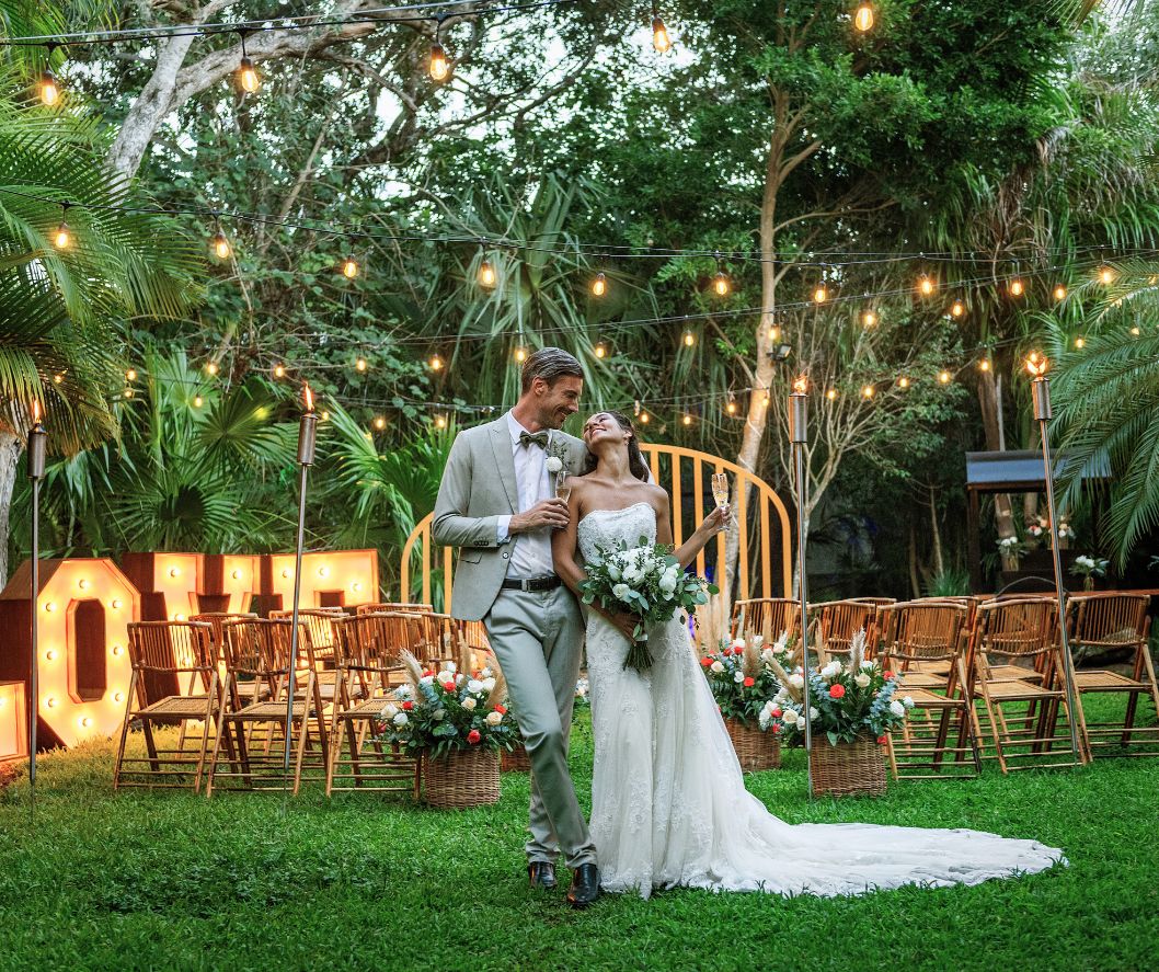 Bride and groom celebrating at an outdoor garden wedding reception with string lights and tropical greenery
