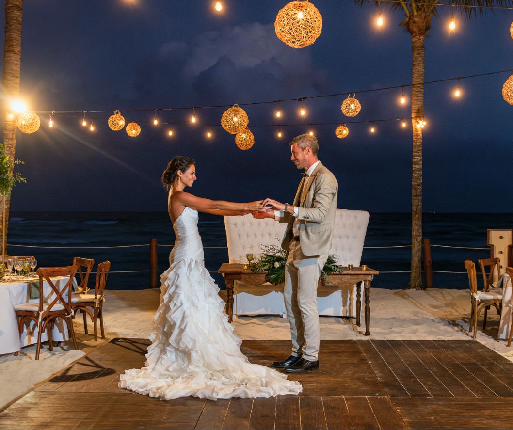 Couple dancing under string lights at a romantic beachfront wedding reception at night