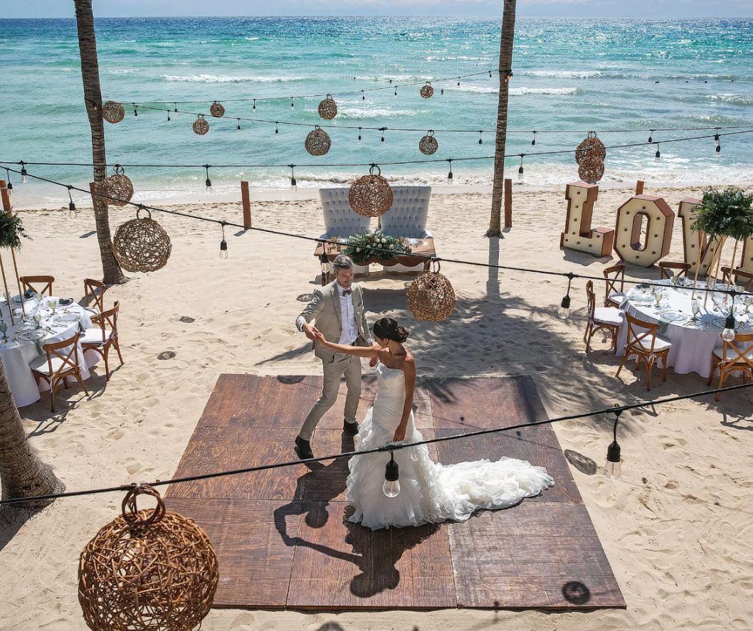 Aerial view of bride and groom dancing on a beachside reception floor surrounded by ocean and décor