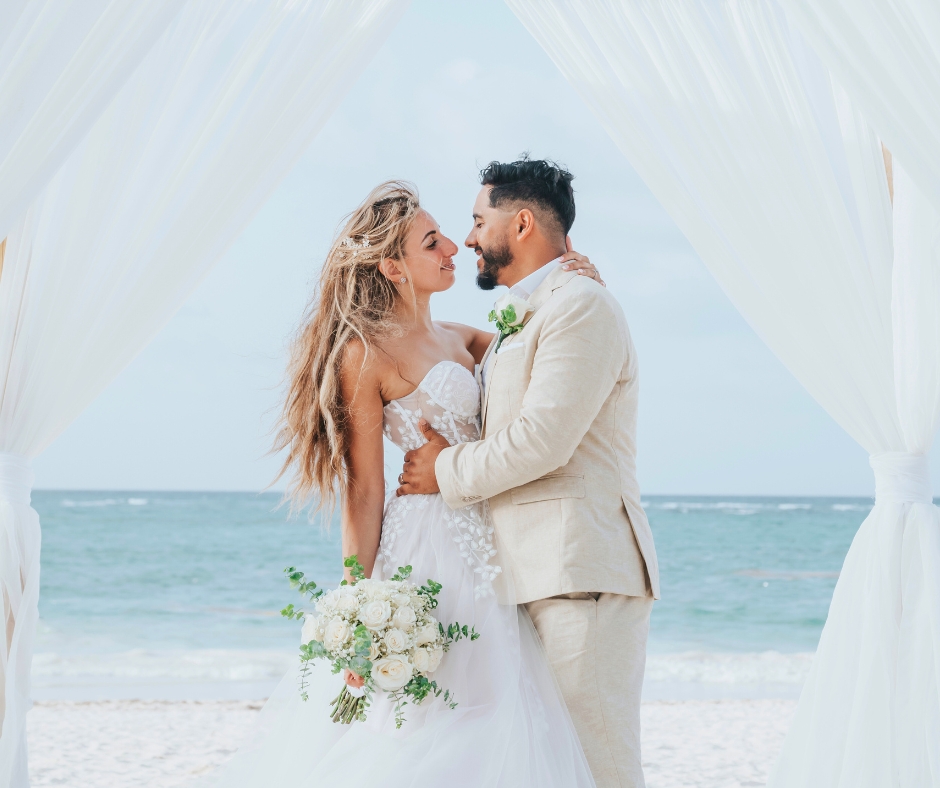 Bride and groom embracing beneath a white canopy during a beachfront wedding ceremony with ocean views