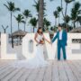 Newlyweds holding hands in front of illuminated “LOVE” letters at a tropical beach wedding venue lined with palm trees.