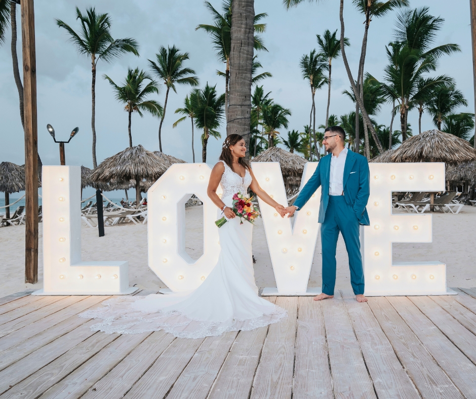Newlyweds holding hands in front of illuminated “LOVE” letters at a tropical beach wedding venue lined with palm trees.