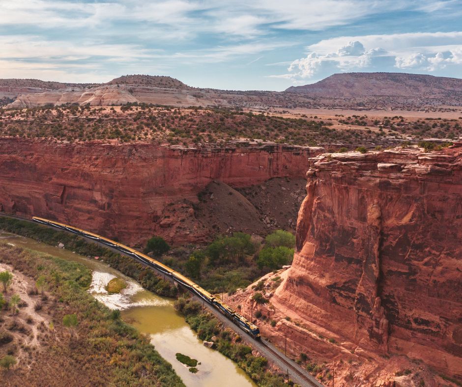 Train traveling along the Colorado River through red rock canyon cliffs in the American Southwest.