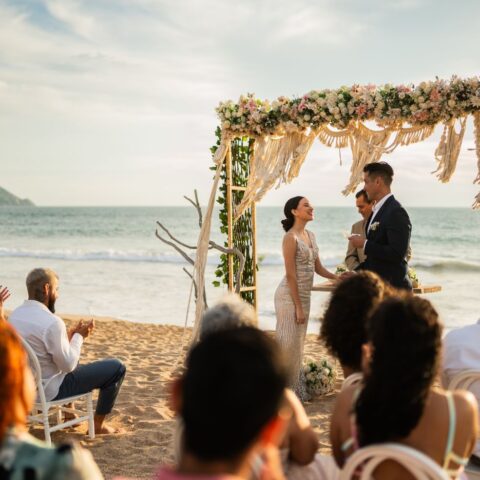 Couple exchanging vows under a floral arch during a beachfront wedding ceremony at sunset.
