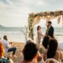 Couple exchanging vows under a floral arch during a beachfront wedding ceremony at sunset.
