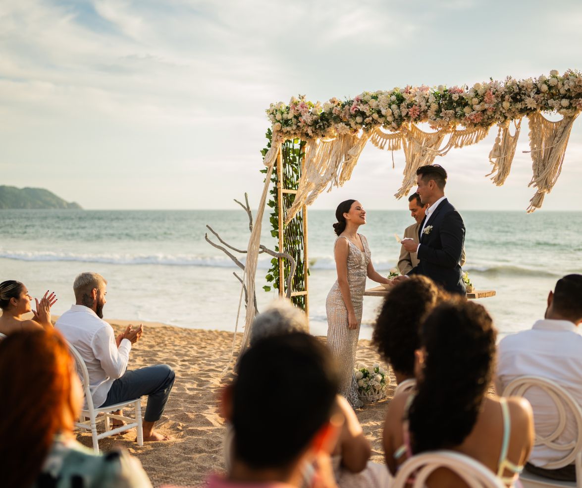 Couple exchanging vows under a floral arch during a beachfront wedding ceremony at sunset.
