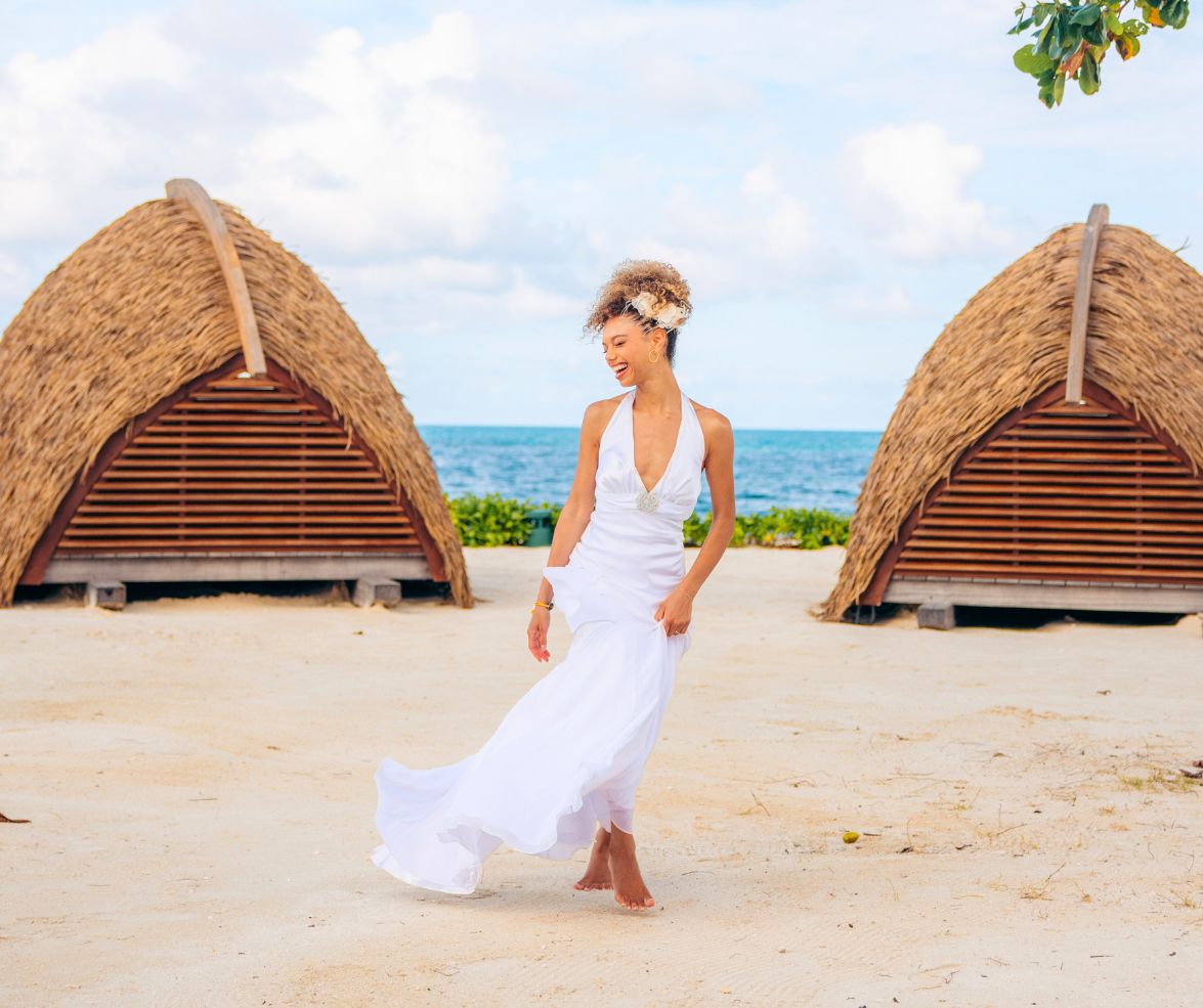 Bride in a flowing white dress walking barefoot on a sandy beach with ocean and cabanas behind her.