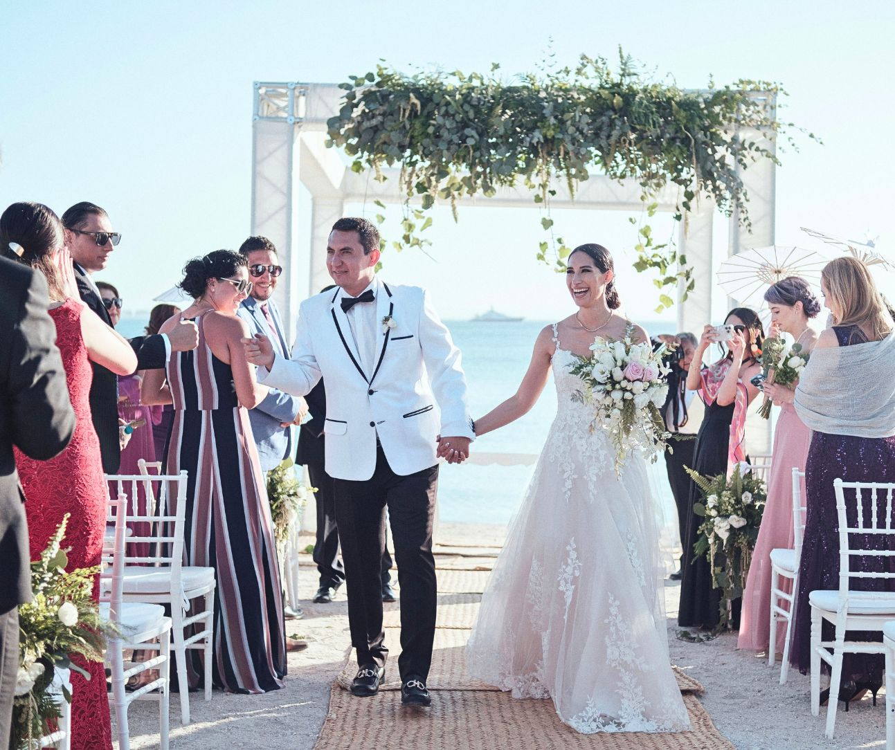 Newlyweds walking down a beach aisle after their ceremony, guests applauding by the ocean.