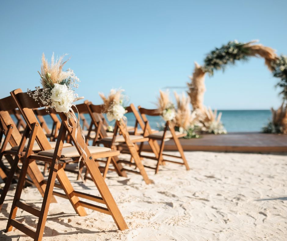 Wooden chairs decorated with floral arrangements set on a sandy beach facing a seaside wedding arch.