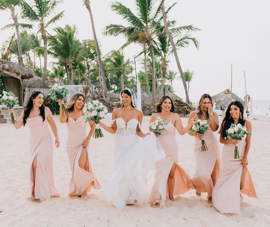 Bride and bridesmaids walking barefoot on a tropical beach, holding bouquets with palm trees behind them.