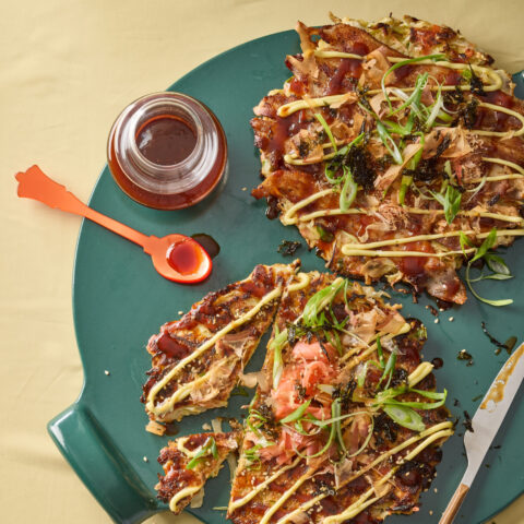 Japanese okonomiyaki pancake cut into wedges on a green plate, topped with scallions, bonito flakes, seaweed, mayonnaise and sweet sauce with a small dish of sauce and spoon beside it.