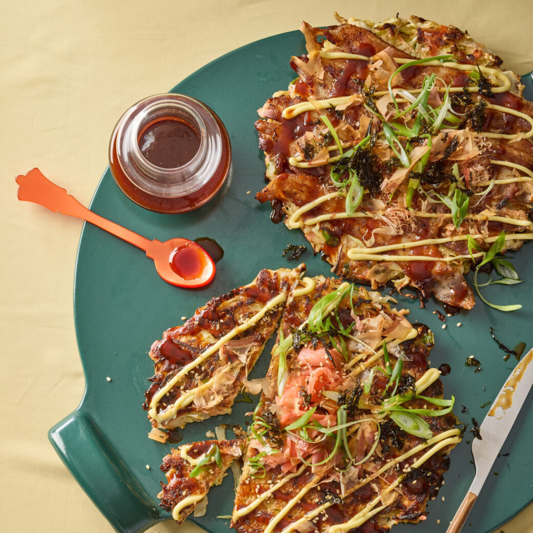 Japanese okonomiyaki pancake cut into wedges on a green plate, topped with scallions, bonito flakes, seaweed, mayonnaise and sweet sauce with a small dish of sauce and spoon beside it.