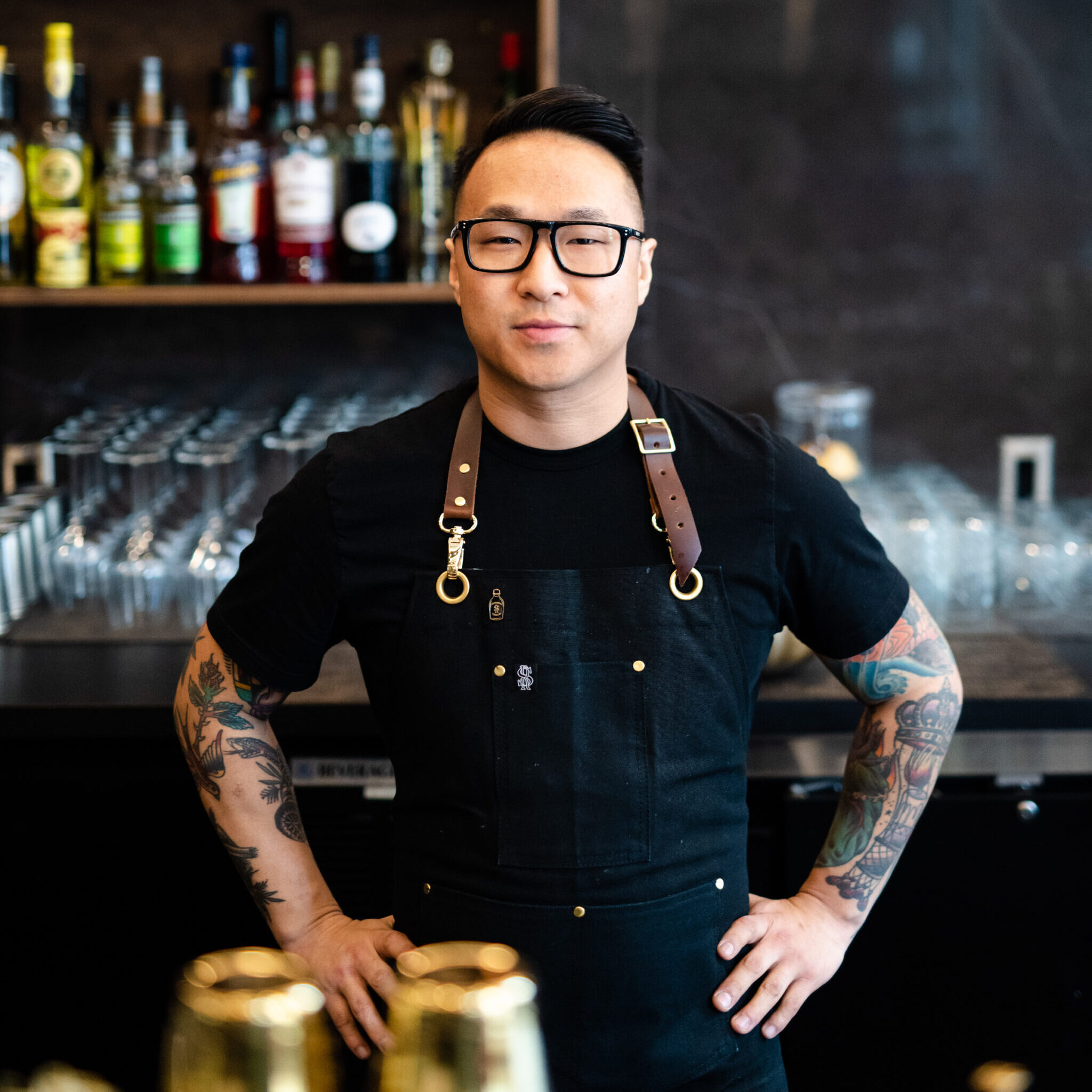 Bespectacled man wearing black tshirt and apron standing behind bar with hands on hips.