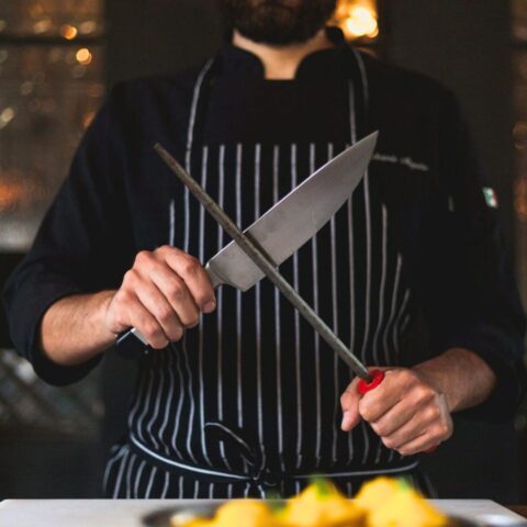 A man in a black chef's jacket and striped apron holding a chef's knife and honing steel.