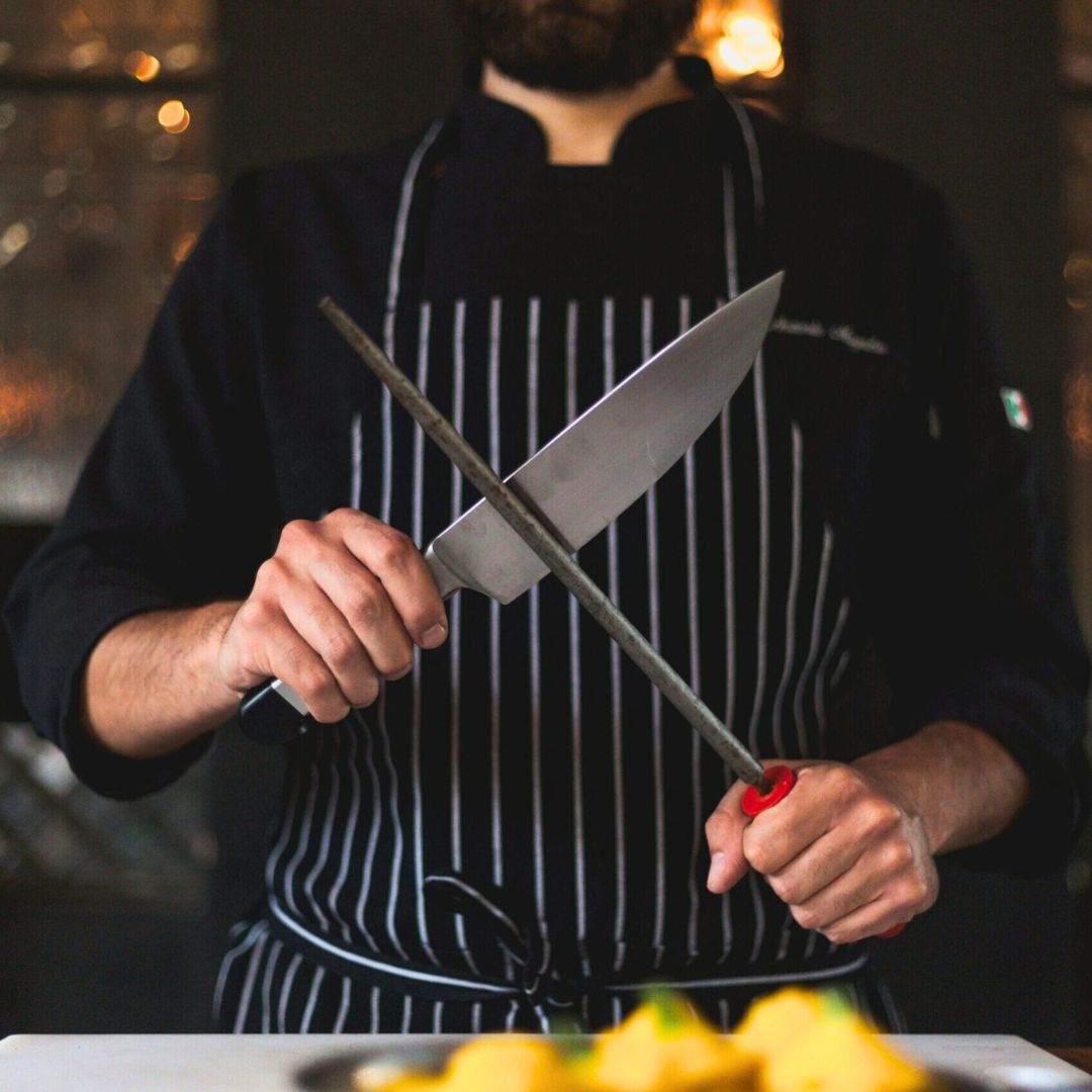 A man in a black chef's jacket and striped apron holding a chef's knife and honing steel.