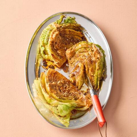 Caramelized cabbage wedges on a platter, drizzled with a creamy sauce and sesame seeds, with a knife resting alongside on a peach-coloured background.