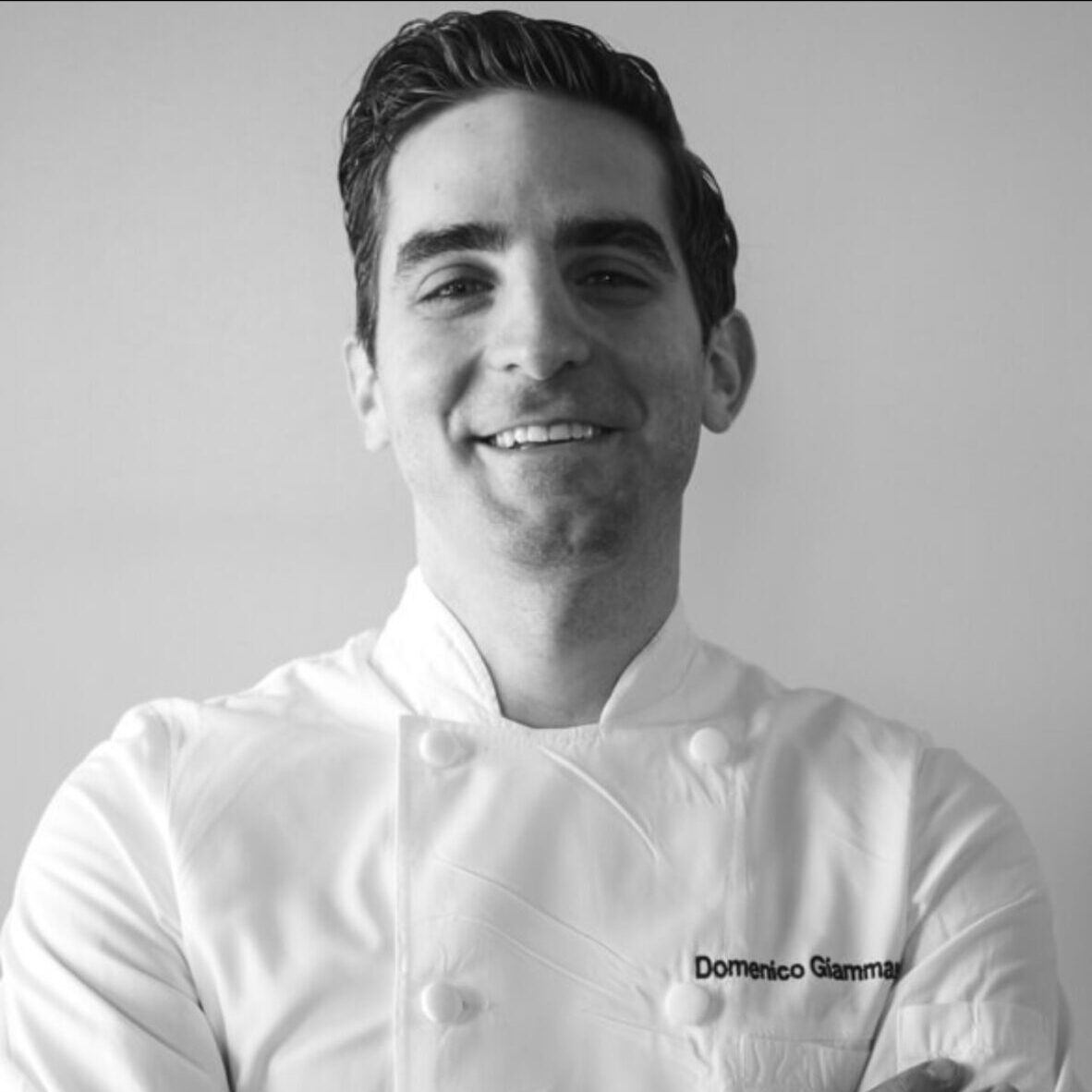 Black and white picture of a smiling man in a chef's jacket.
