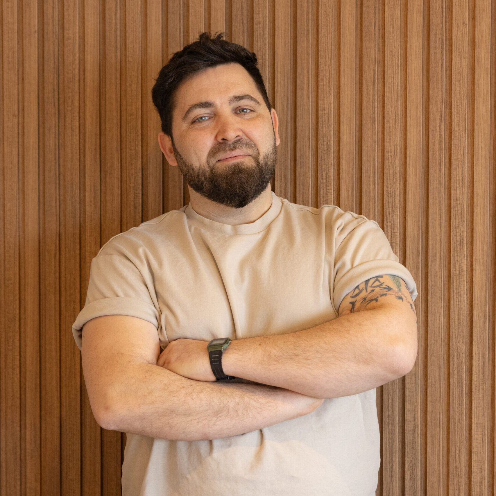 Man with brown hair and beard stands with arms crossed in front of a wood panelled wall.
