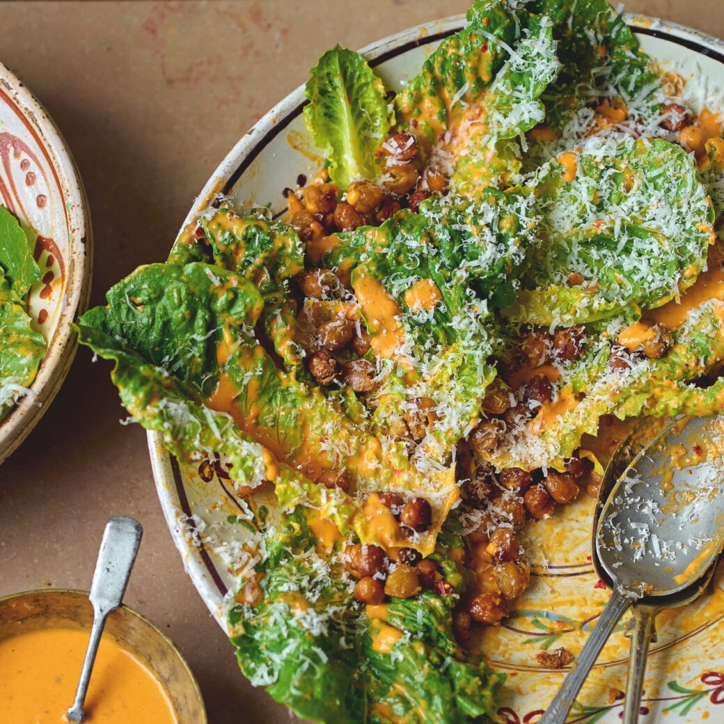 Romaine lettuce tossed with creamy orange dressing and crispy spiced chickpeas, topped with grated cheese, served on a plate with spoons and extra dressing on the side.