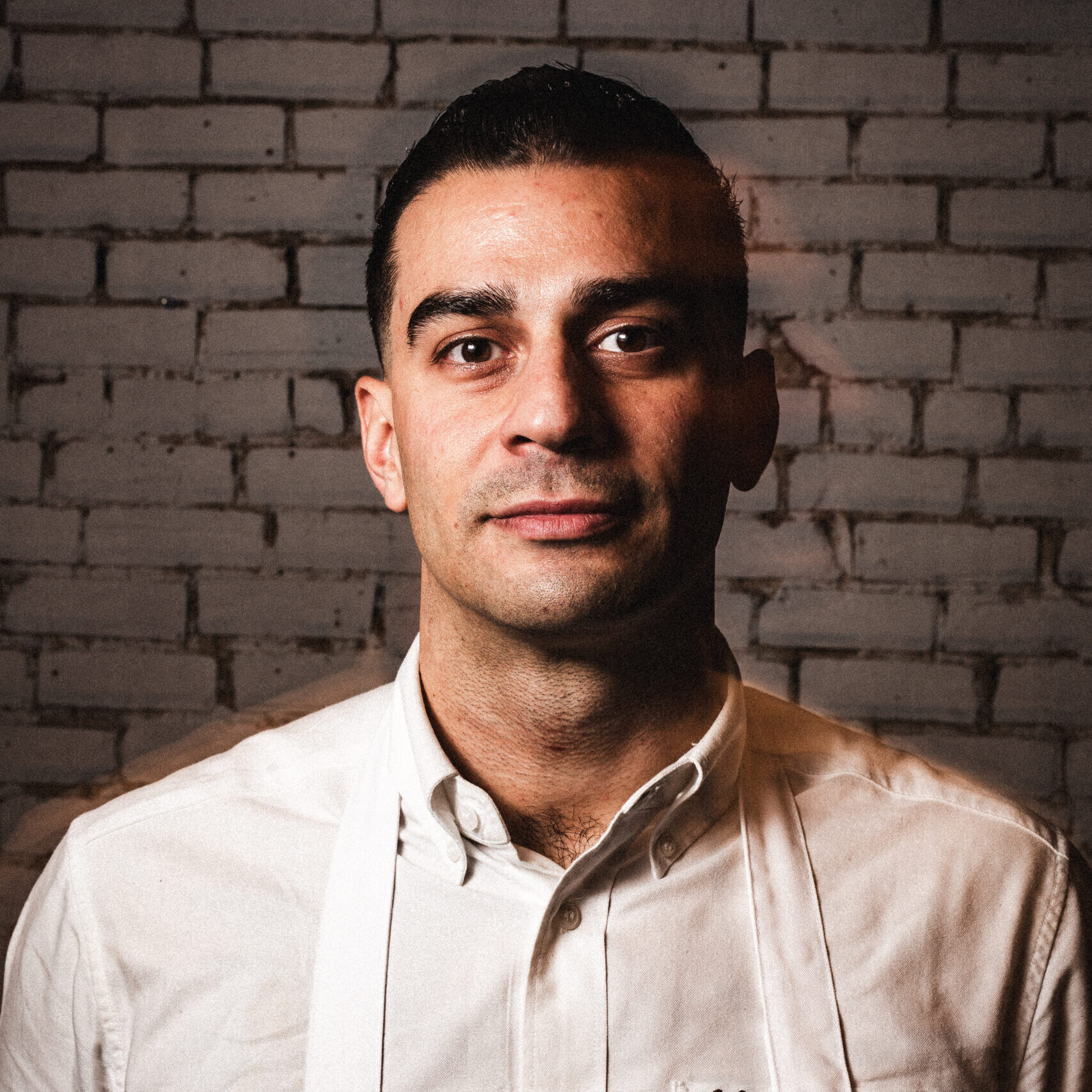 Close-up of a man with black hair wearing a white button up and white apron in front of a white brick wall. 