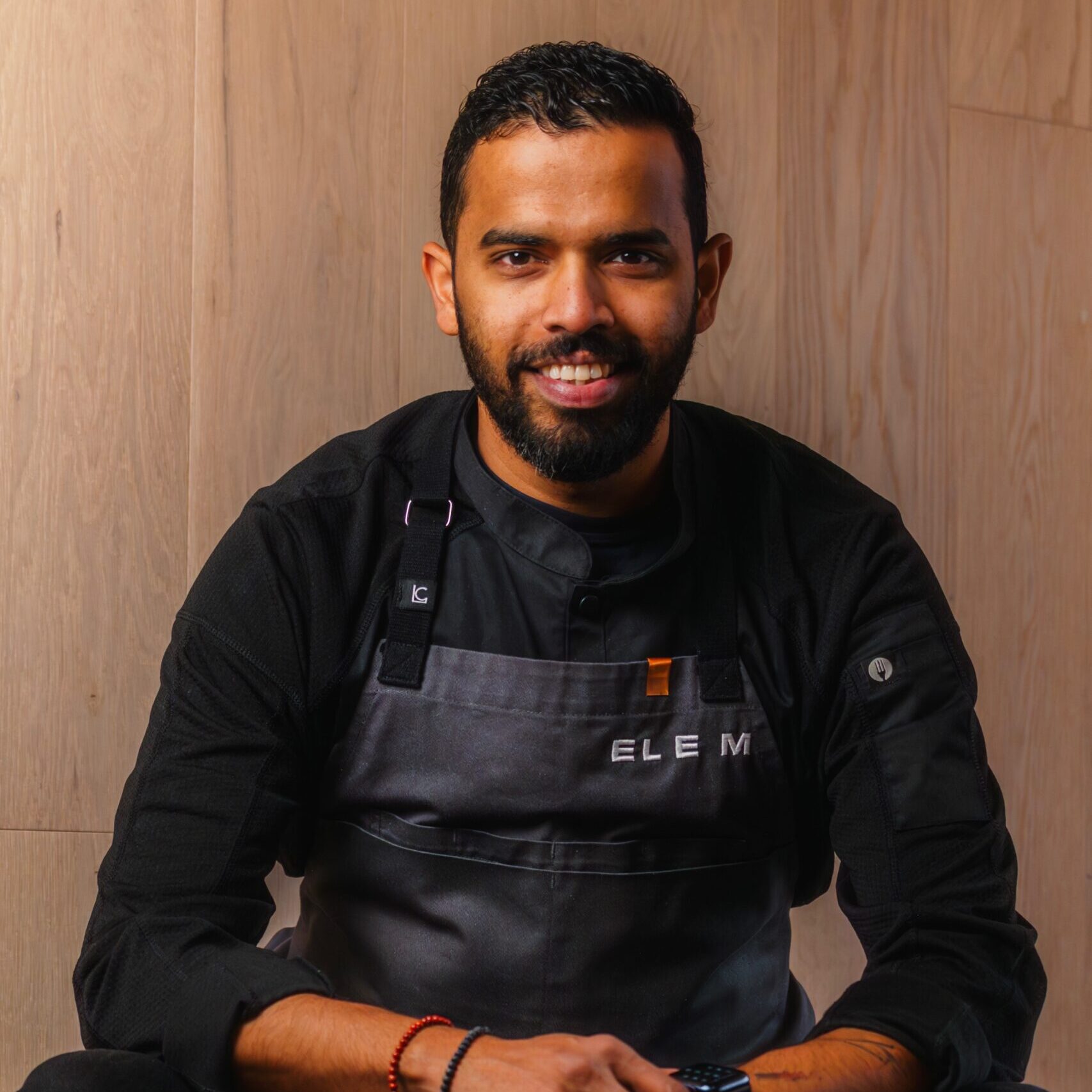 Smiling man in a black shirt and black apron sitting in front of a wooden wall. 