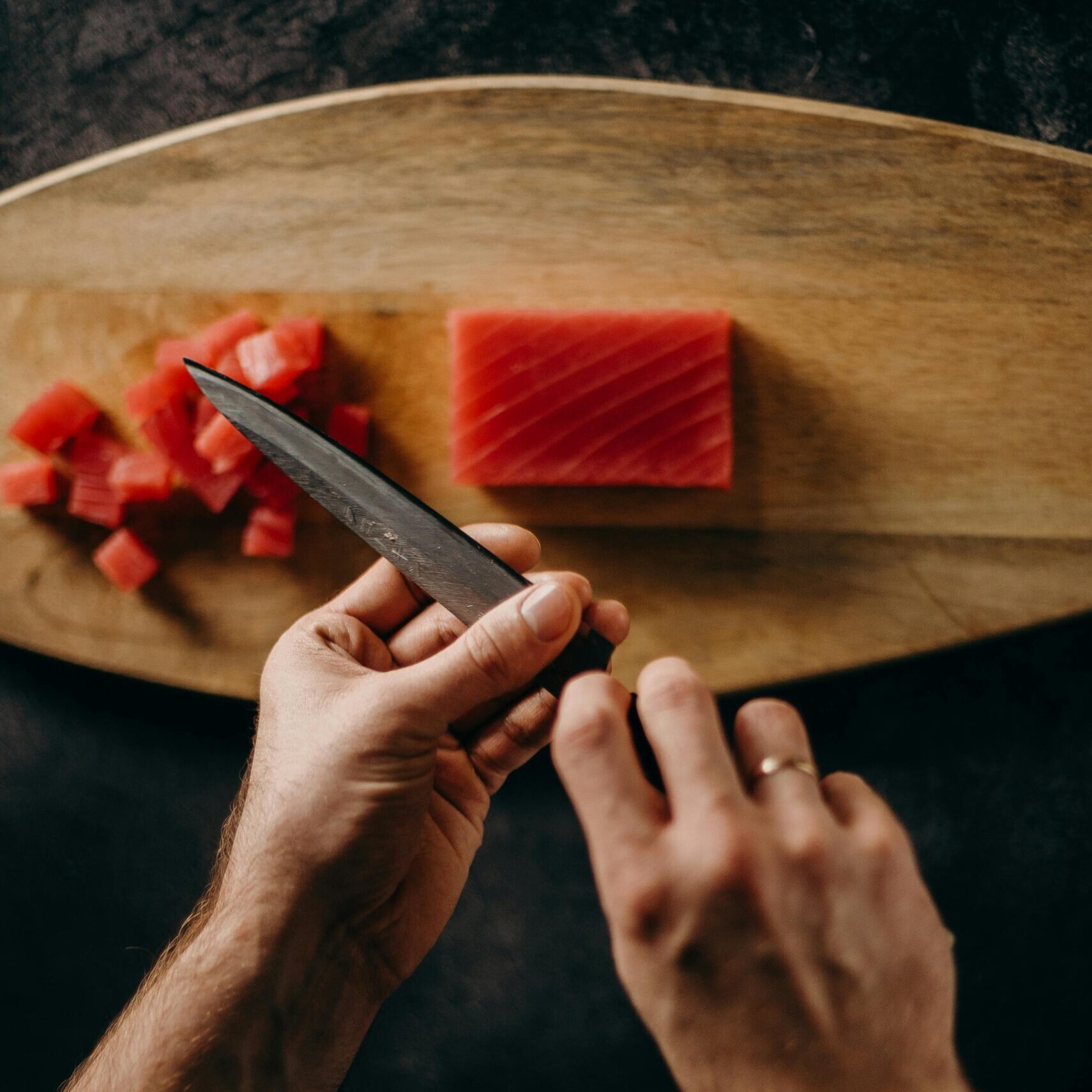 Two hands holding a knife above a wooden cutting board with raw tuna.