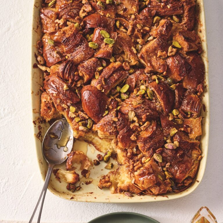 Baked French toast topped with chopped nuts and syrup in a dish, with a serving on a plate and a small pitcher of syrup alongside.