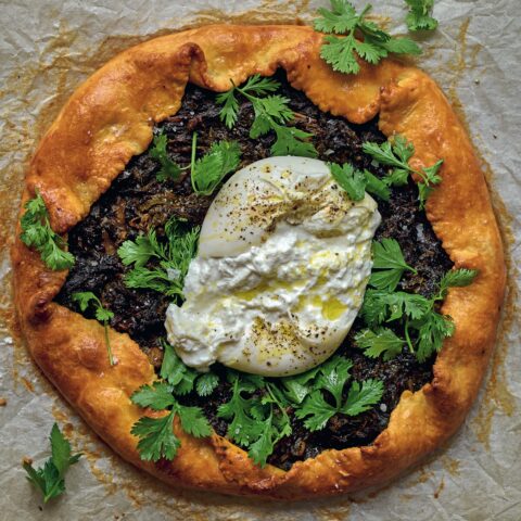Rustic savoury galette filled with greens, topped with fresh herbs and a ball of burrata, set on parchment paper.