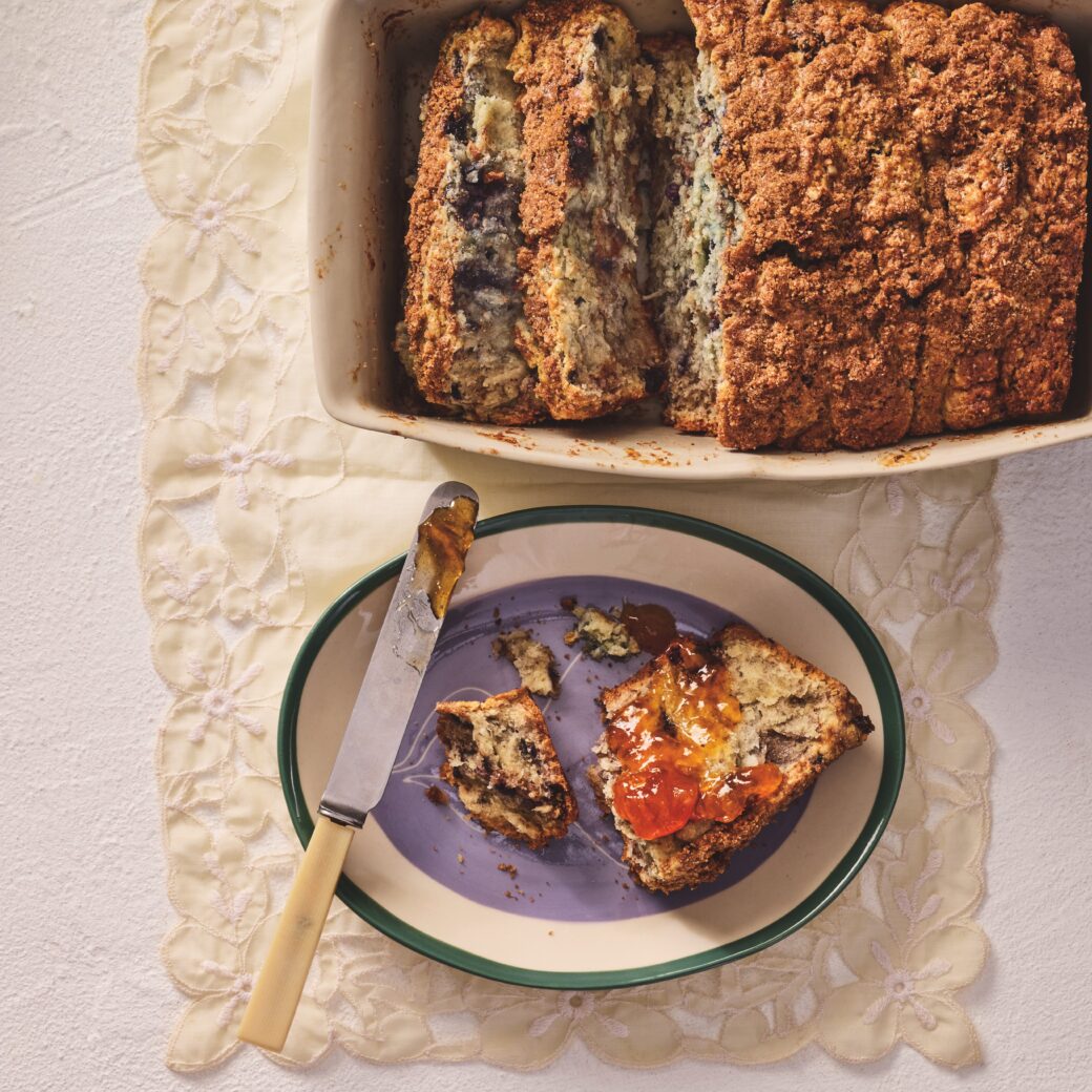 Blueberry pull-apart loaf sliced in a baking dish, with a piece spread with jam on a plate and a butter knife alongside.