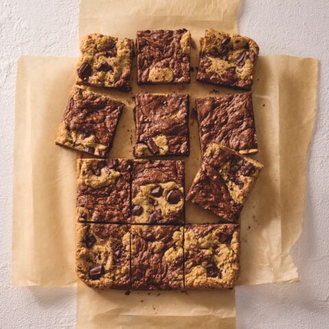 Chocolate chip brownie-cookie bars (brookies) cut into squares and arranged on parchment paper, showing a marbled brownie and cookie dough top with chunks of chocolate.