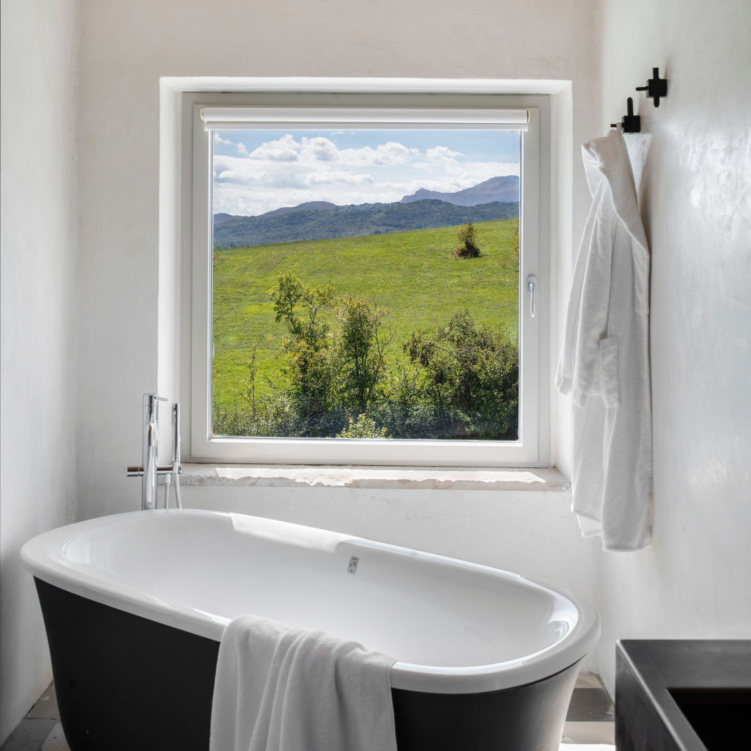 A black bathtub in a white bathroom with a square window overlooking greenery. 