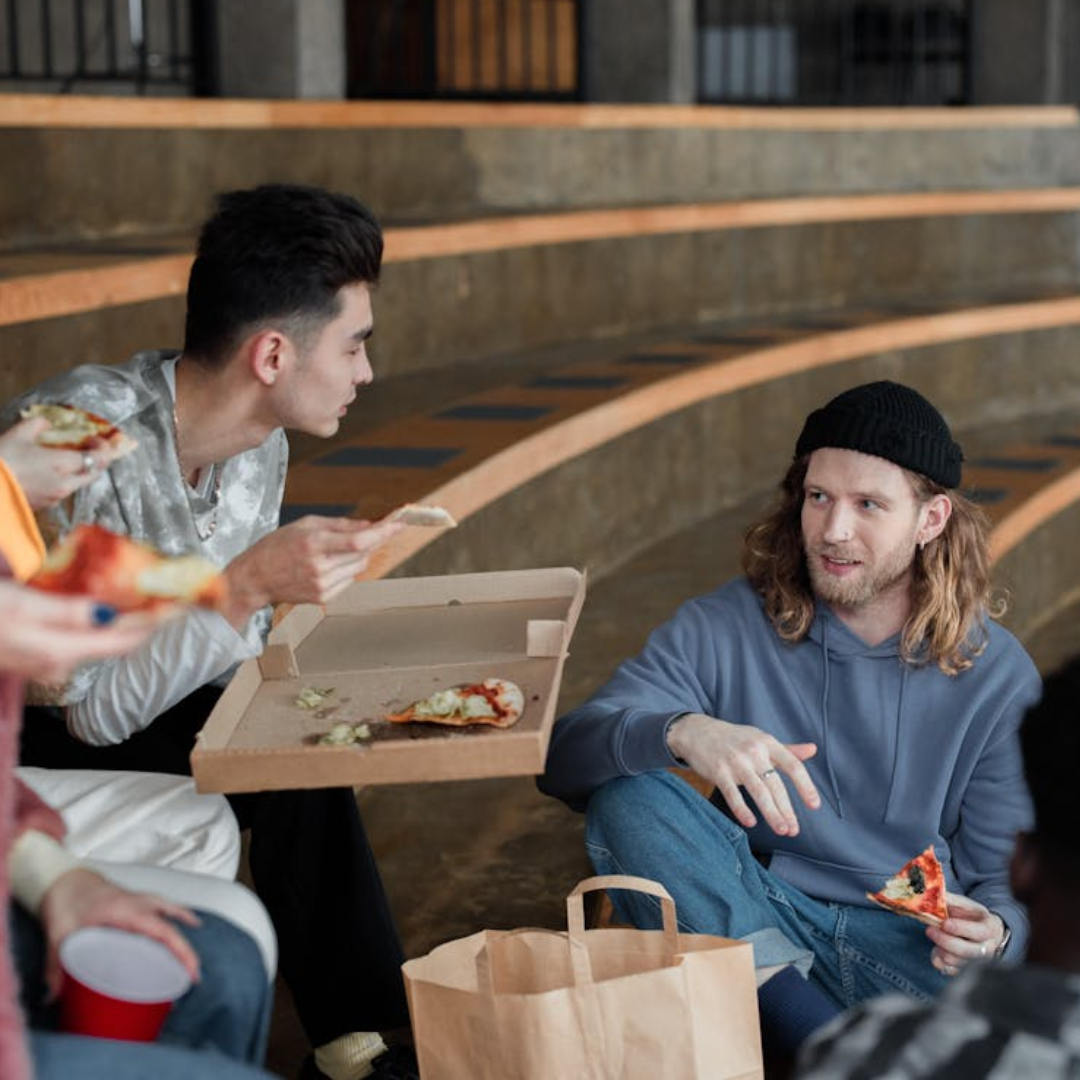 Two men speaking while eating pizza on steps. 