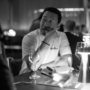 Black and white image of a man in a chef's jacket sitting at the table resting his thumb against his lips.