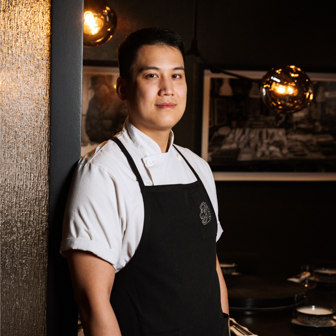 Man in white chef's coat and black apron smiling and leaning against a doorframe.