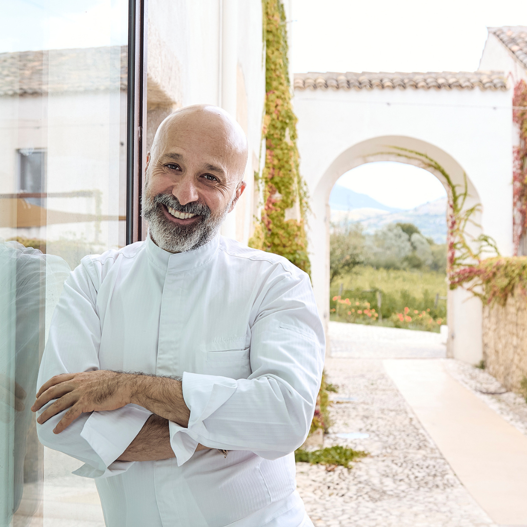 A bald bearded man smiling in a white chef's jacket with arms crossed leaning against a glass window. 