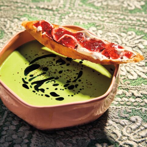 Chilled green pea soup drizzled with basil oil in a bowl, served with a toasted baguette slice topped with creamy spread and strawberry jam.
