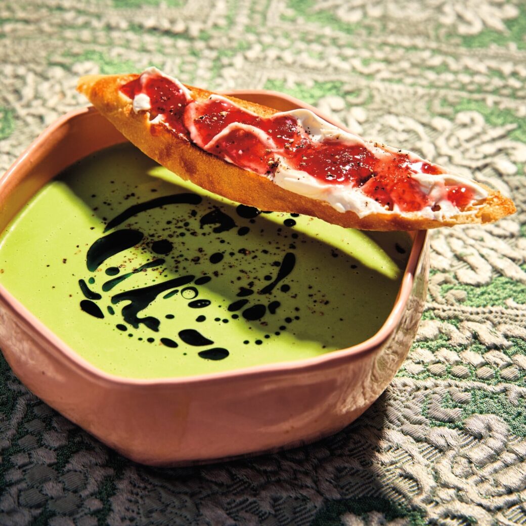 Chilled green pea soup drizzled with basil oil in a bowl, served with a toasted baguette slice topped with creamy spread and strawberry jam.
