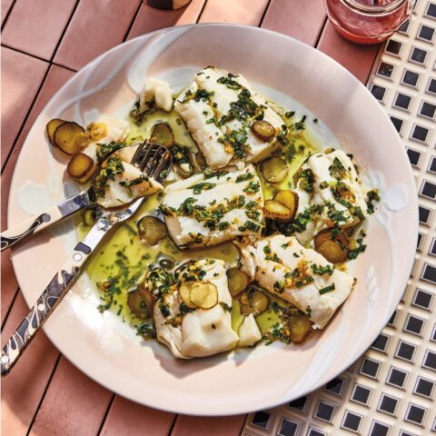 Poached cod fillets topped with a scallion herb sauce and sliced pickles, served on a plate with utensils, alongside drinks on a tiled table.