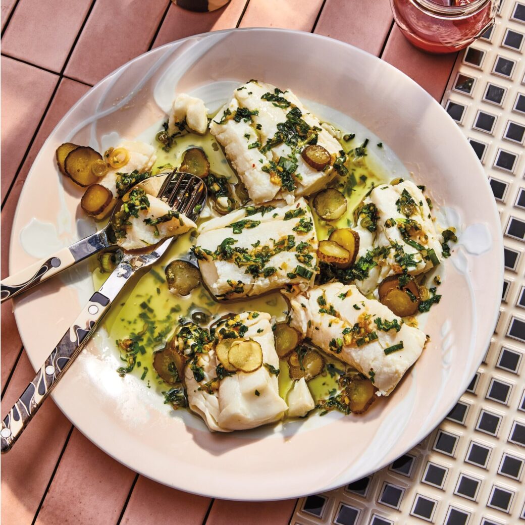 Poached cod fillets topped with a scallion herb sauce and sliced pickles, served on a plate with utensils, alongside drinks on a tiled table.