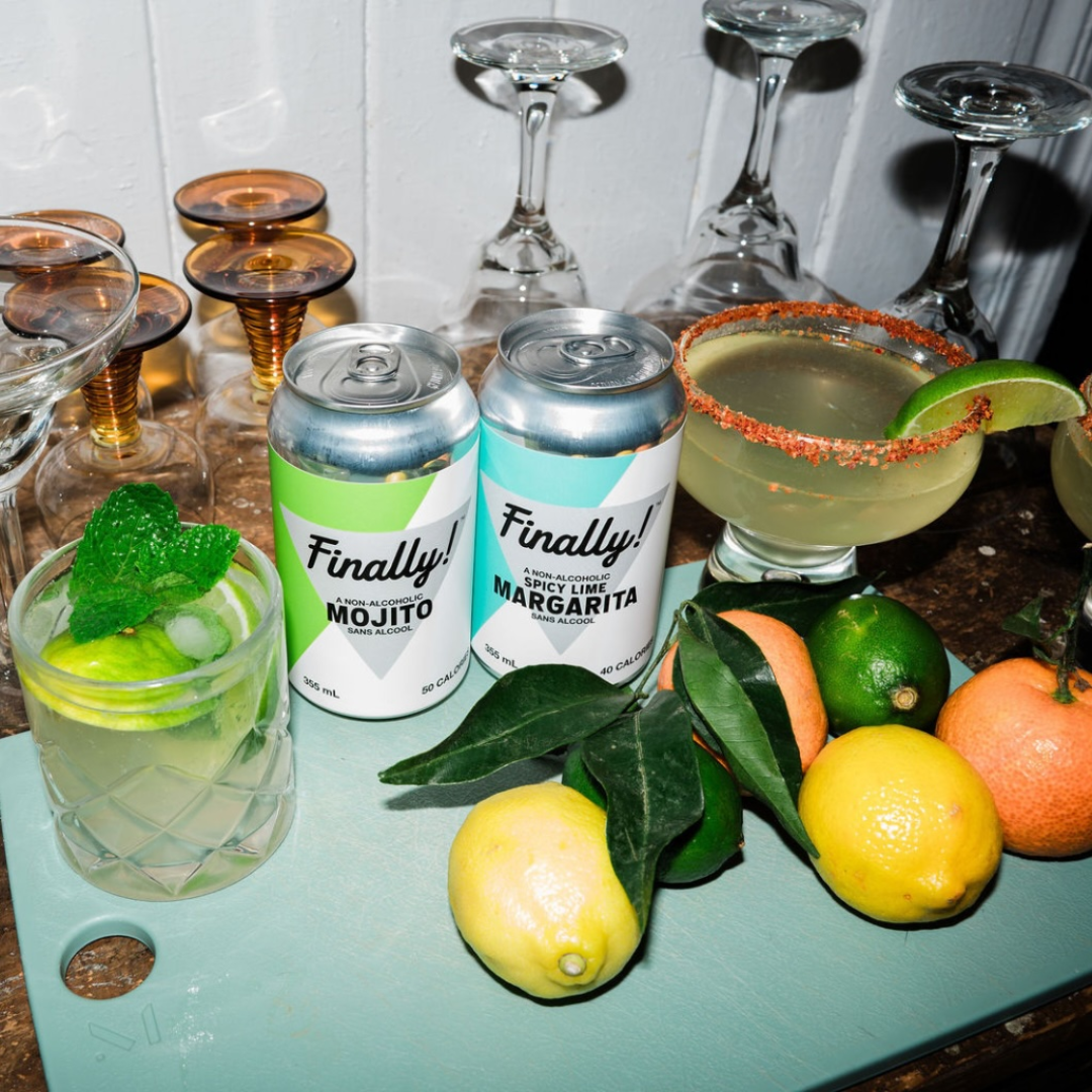Two cans in between cocktails, lemons, oranges and upside-down glasses on a green cutting board.