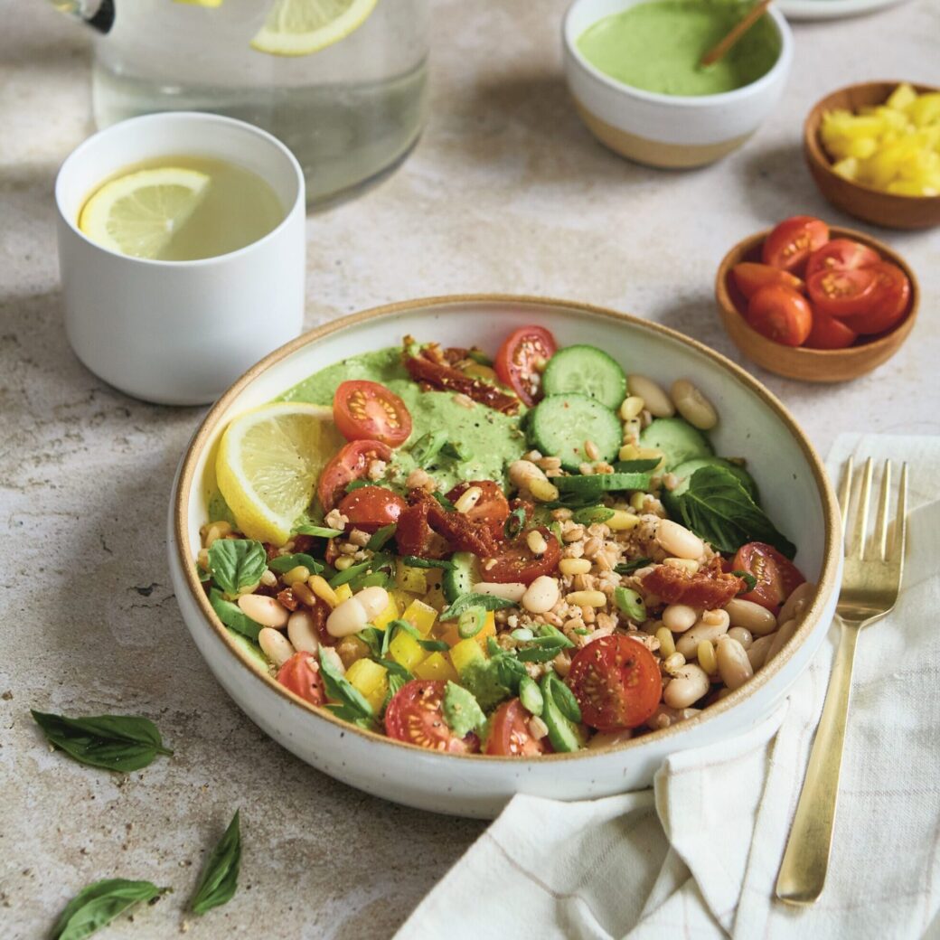 Grain and bean bowl topped with cherry tomatoes, cucumber, white beans, herbs and a creamy green pesto dressing, served with lemon water and fresh ingredients on the side.