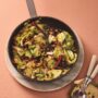 Cast-iron skillet filled with hot and sour cabbage stir-fry, tossed with dried chilies, peanuts and scallions, set on a soft peach background with serving spoons nearby.