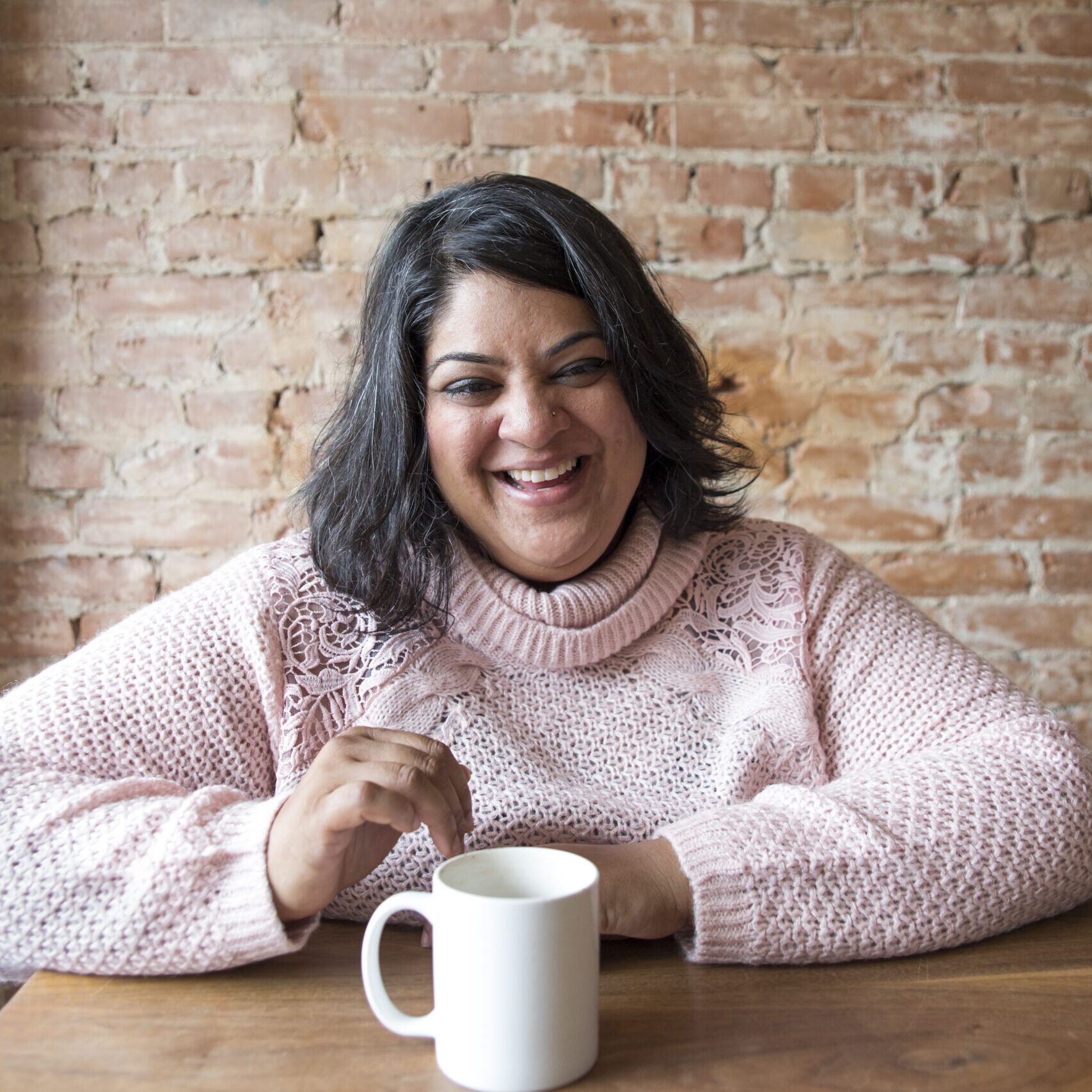 Woman in pink sweater smiling at a table with a white coffee mug. 