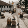A man in a beekeeping suit stands with one leg on a barrel and one arm raised holding a bottle of yellow liqueur.