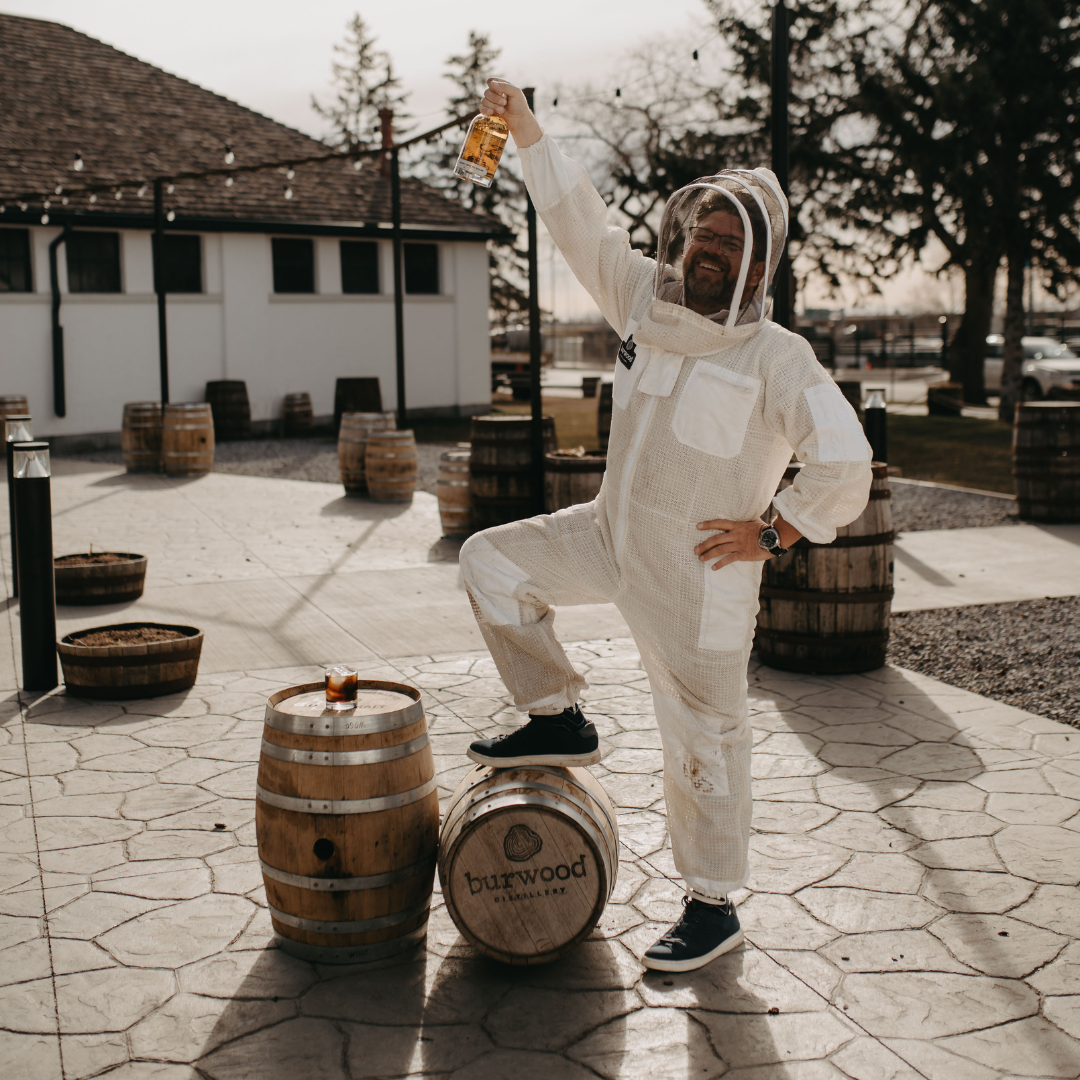 A man in a beekeeping suit stands with one leg on a barrel and one arm raised holding a bottle of yellow liqueur.