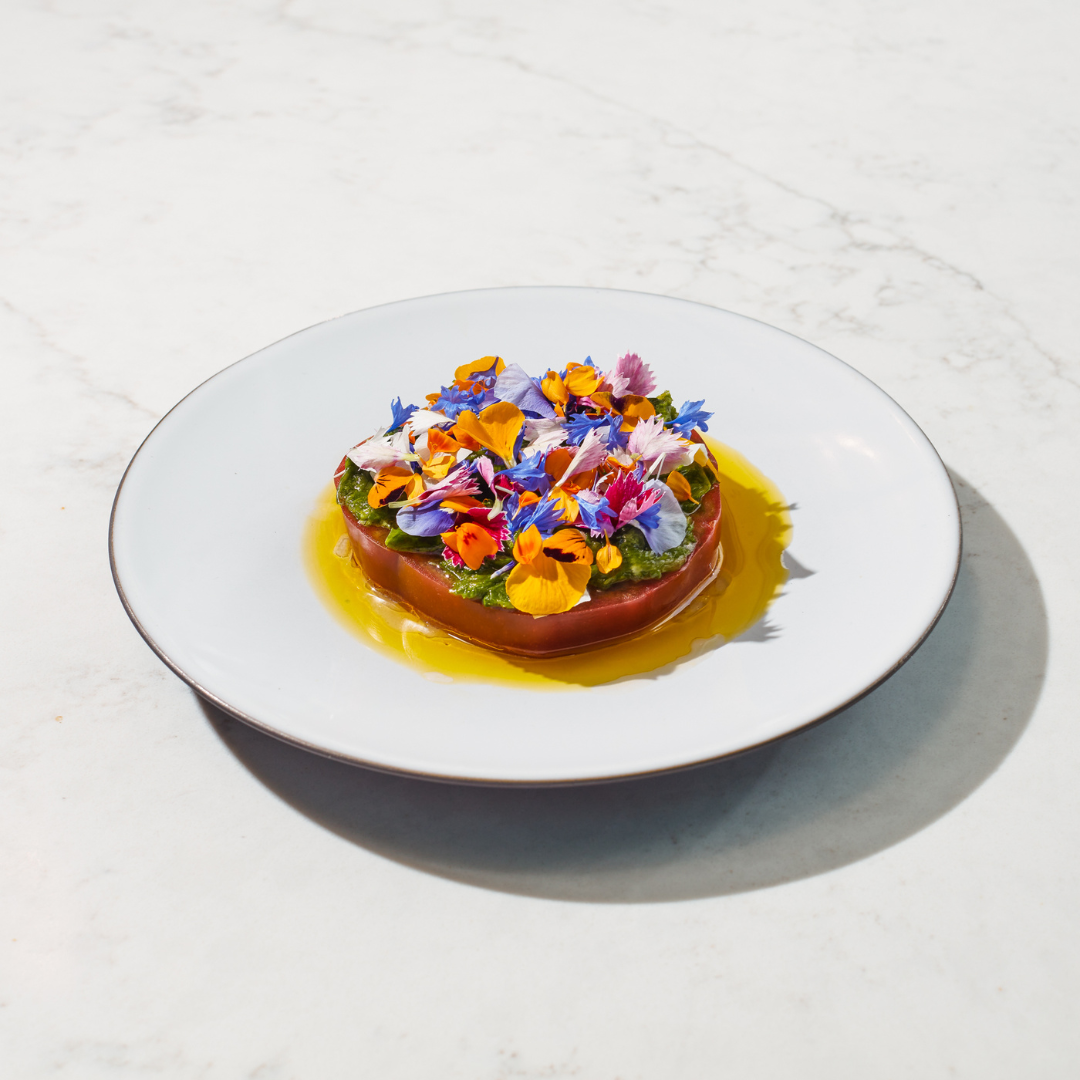 Tomato steak in yellow sauce covered in edible flowers on a white plate. 