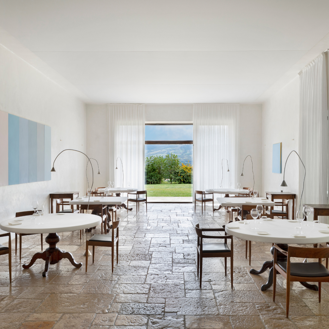 A dining room with white walls, white tables, wooden chairs and a large window at the back wall overlooking greenery.