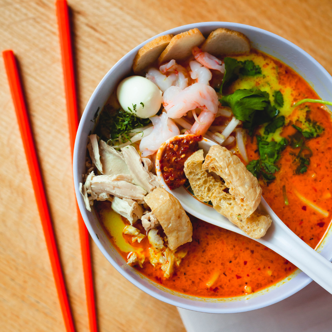Bowl of laksa soup on a wooden table with a pair of orange chopsticks.